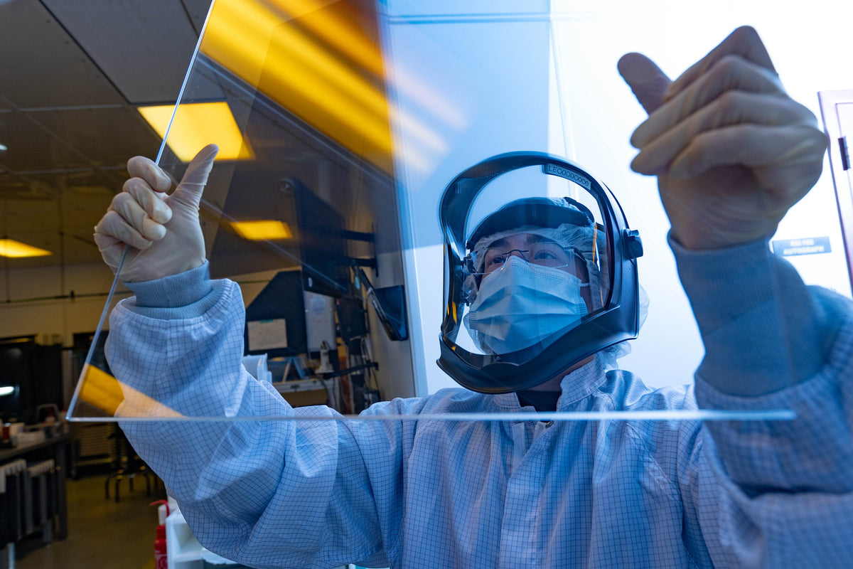 A person in a cleanroom protective suit and face shield holds up a large transparent glass panel in a laboratory setting.
